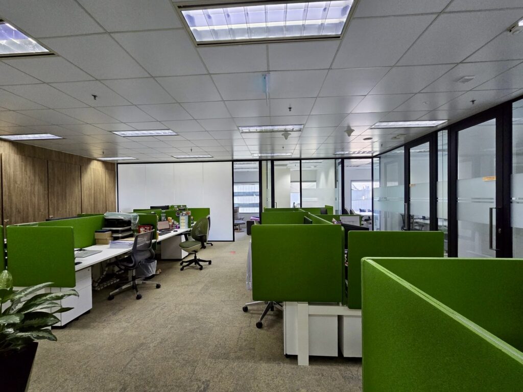 Open-plan office with green partitioned desks, chairs, computers, and large windows. The lights are on, but the space appears mostly unoccupied.