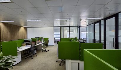 Open-plan office with green partitioned desks, chairs, computers, and large windows. The lights are on, but the space appears mostly unoccupied.