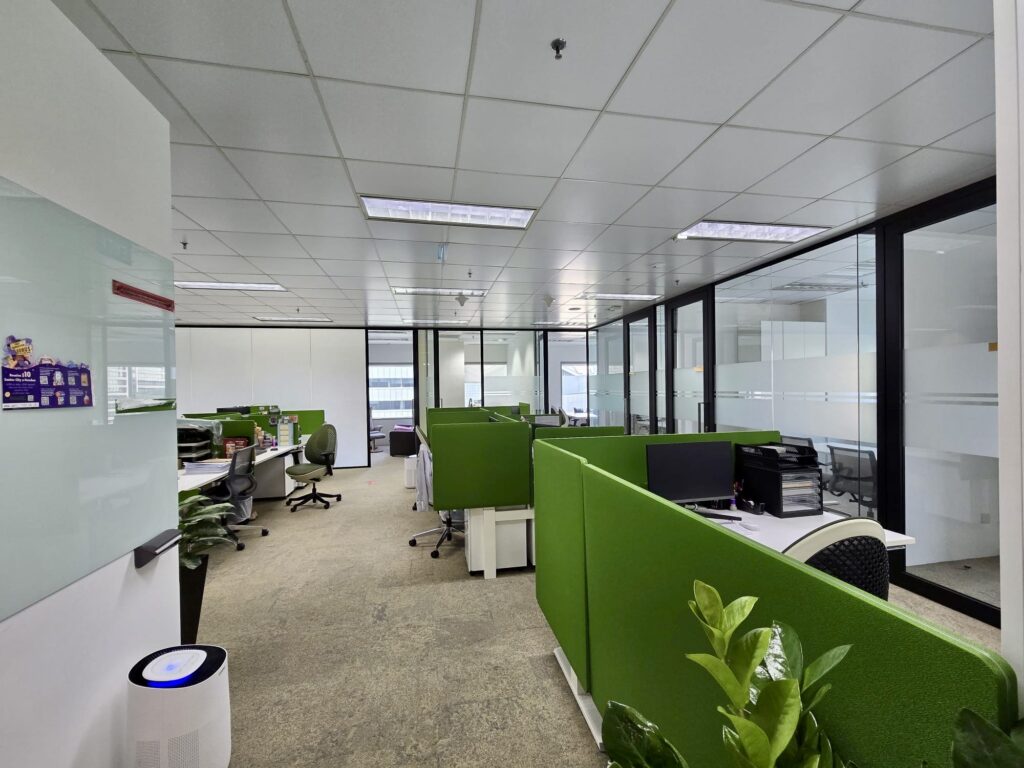 Modern office with green partitioned desks, computers, office chairs, glass-walled meeting rooms, whiteboards, and indoor plants under fluorescent lighting.