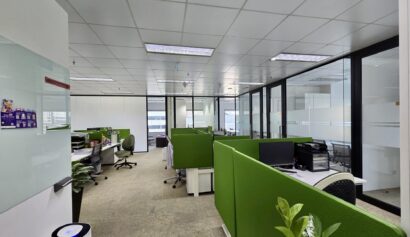 Modern office with green partitioned desks, computers, office chairs, glass-walled meeting rooms, whiteboards, and indoor plants under fluorescent lighting.