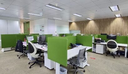 Modern open-plan office with green partitioned desks, office chairs, computers, and overhead fluorescent lighting. The workspace appears empty with no people present.