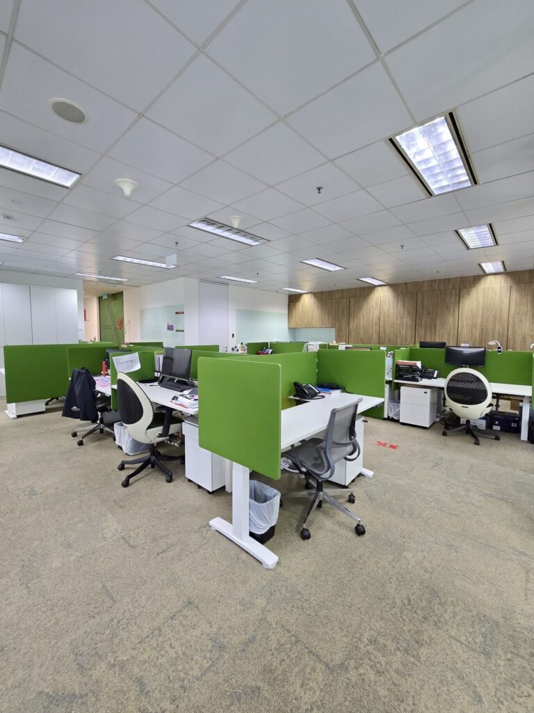 Modern open-plan office with green partitioned desks, office chairs, computers, and overhead fluorescent lighting. The workspace appears empty with no people present.