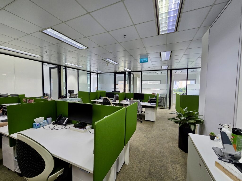 Modern open-plan office with green partitioned desks, computers, chairs, and office supplies. Large windows let in natural light. Some plants and personal items are visible.