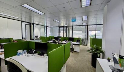 Modern open-plan office with green partitioned desks, computers, chairs, and office supplies. Large windows let in natural light. Some plants and personal items are visible.