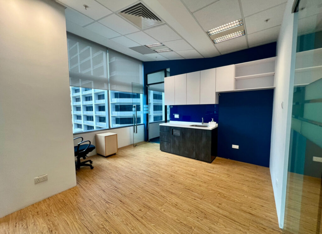 Modern office space with wooden flooring, a desk and chair near large windows, and a kitchenette area with cupboards against a blue feature wall.