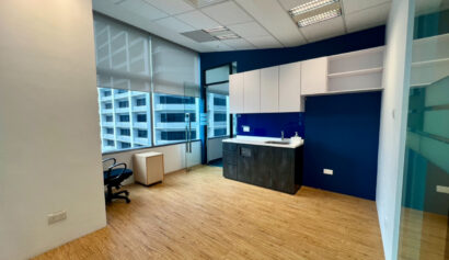 Modern office space with wooden flooring, a desk and chair near large windows, and a kitchenette area with cupboards against a blue feature wall.
