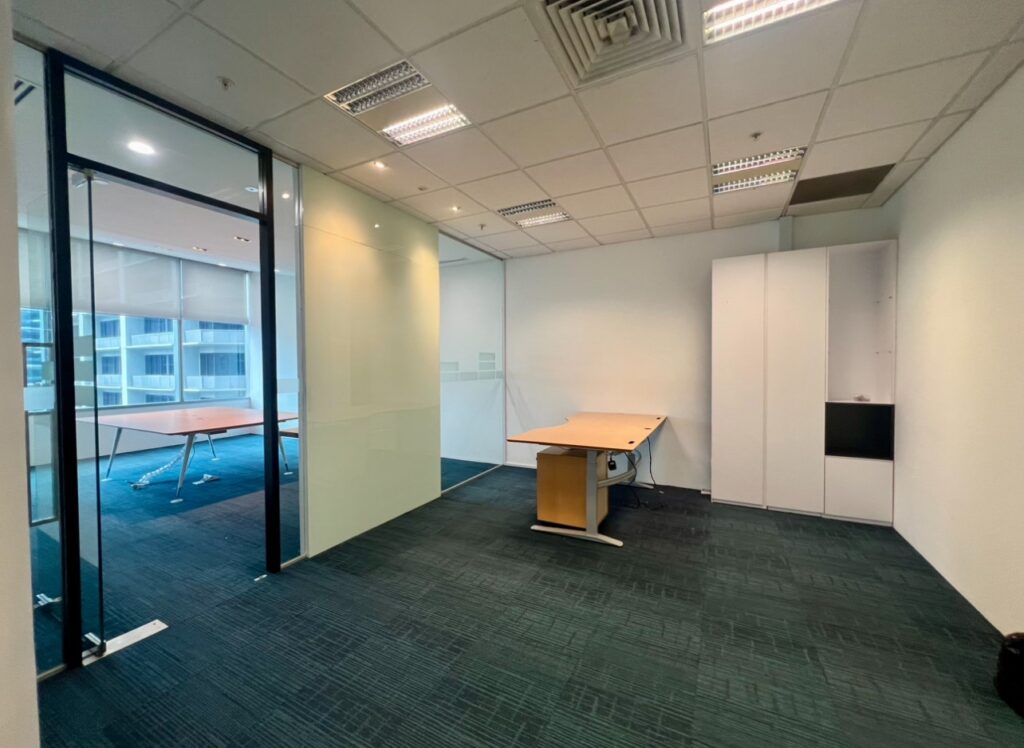 An empty office with two desks, glass partition, built-in storage, and overhead fluorescent lighting. Floor-to-ceiling windows are visible in the adjoining room.