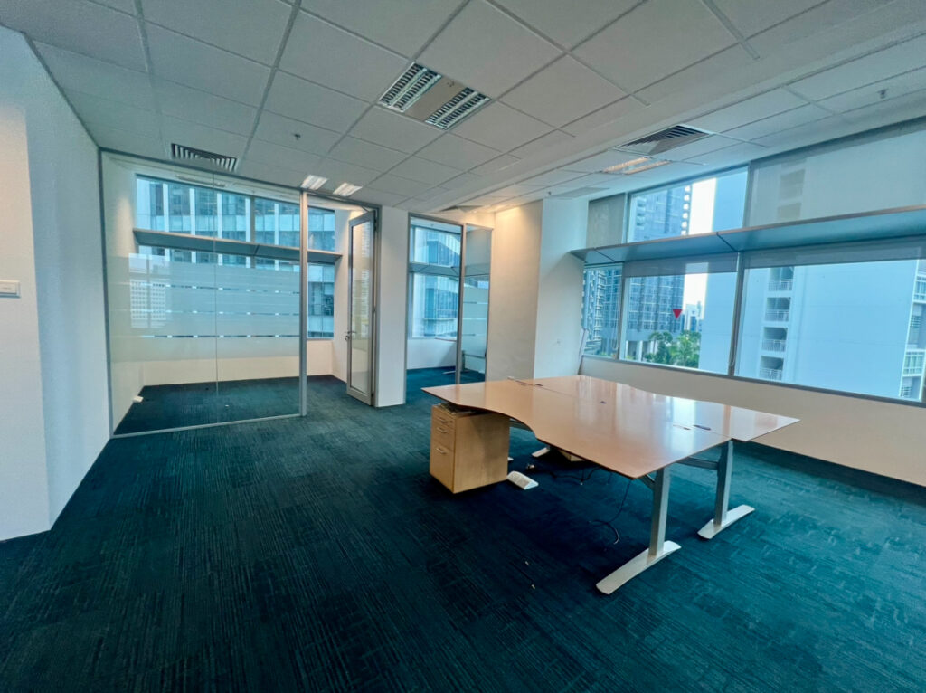 A modern empty office with blue carpet, a large desk, glass partitions, and windows overlooking tall city buildings.