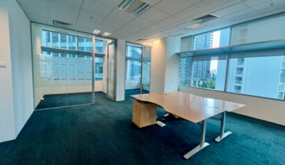 A modern empty office with blue carpet, a large desk, glass partitions, and windows overlooking tall city buildings.