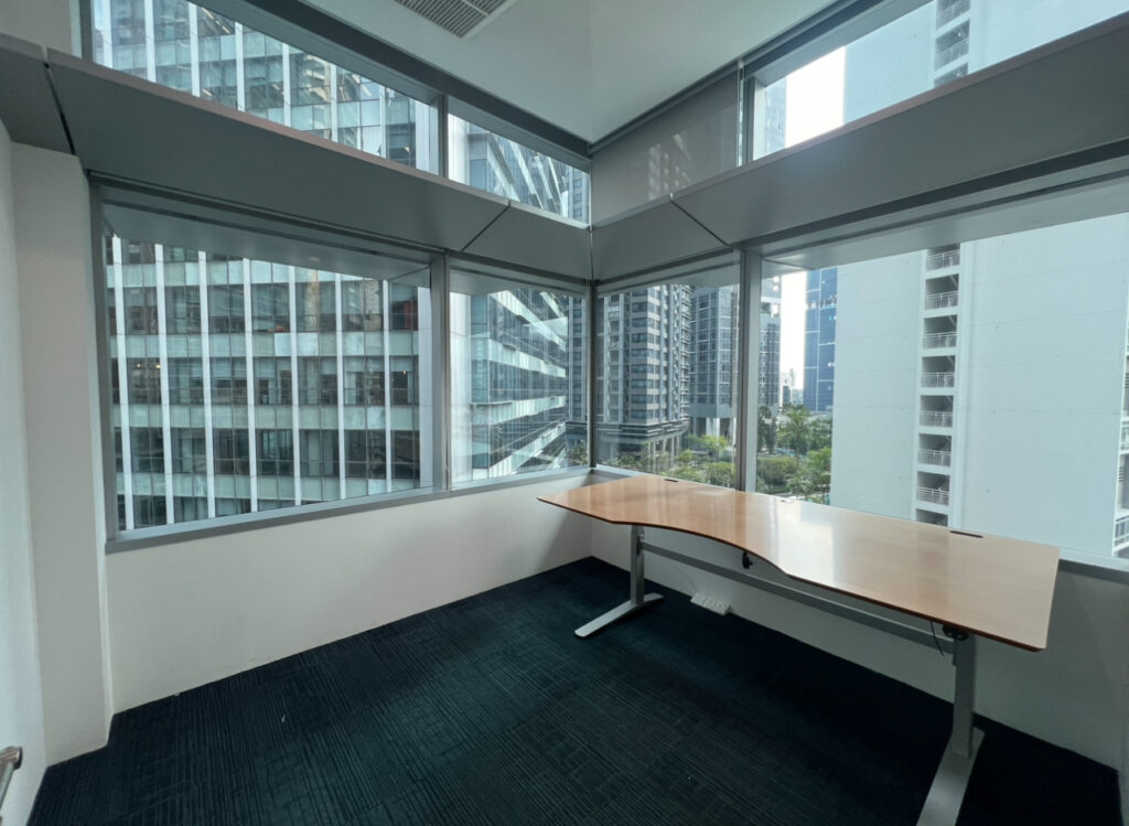 A corner office with large windows, an empty wooden desk, and a view of modern high-rise buildings outside.