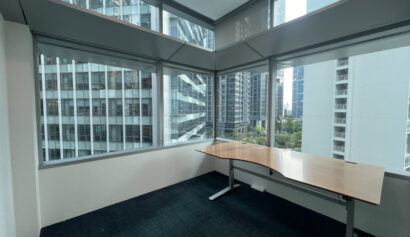 A corner office with large windows, an empty wooden desk, and a view of modern high-rise buildings outside.