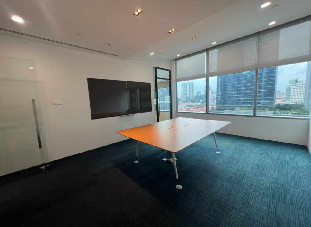 A modern conference room with a large table, wall-mounted screen, glass door, and floor-to-ceiling windows overlooking city buildings.