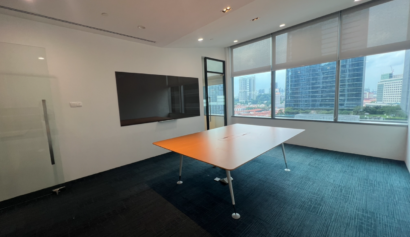 A modern conference room with a large table, wall-mounted screen, glass door, and floor-to-ceiling windows overlooking city buildings.