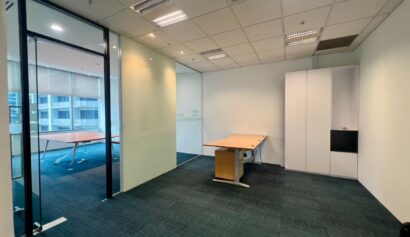 An empty office with two desks, glass partition, built-in storage, and overhead fluorescent lighting. Floor-to-ceiling windows are visible in the adjoining room.