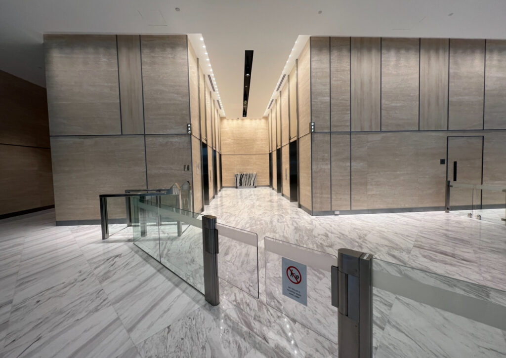 A modern building lobby with marble floors, tall beige walls, and multiple lifts at the far end, separated by glass barriers with a no-smoking sign.