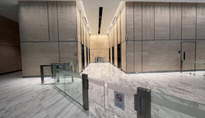 A modern building lobby with marble floors, tall beige walls, and multiple lifts at the far end, separated by glass barriers with a no-smoking sign.