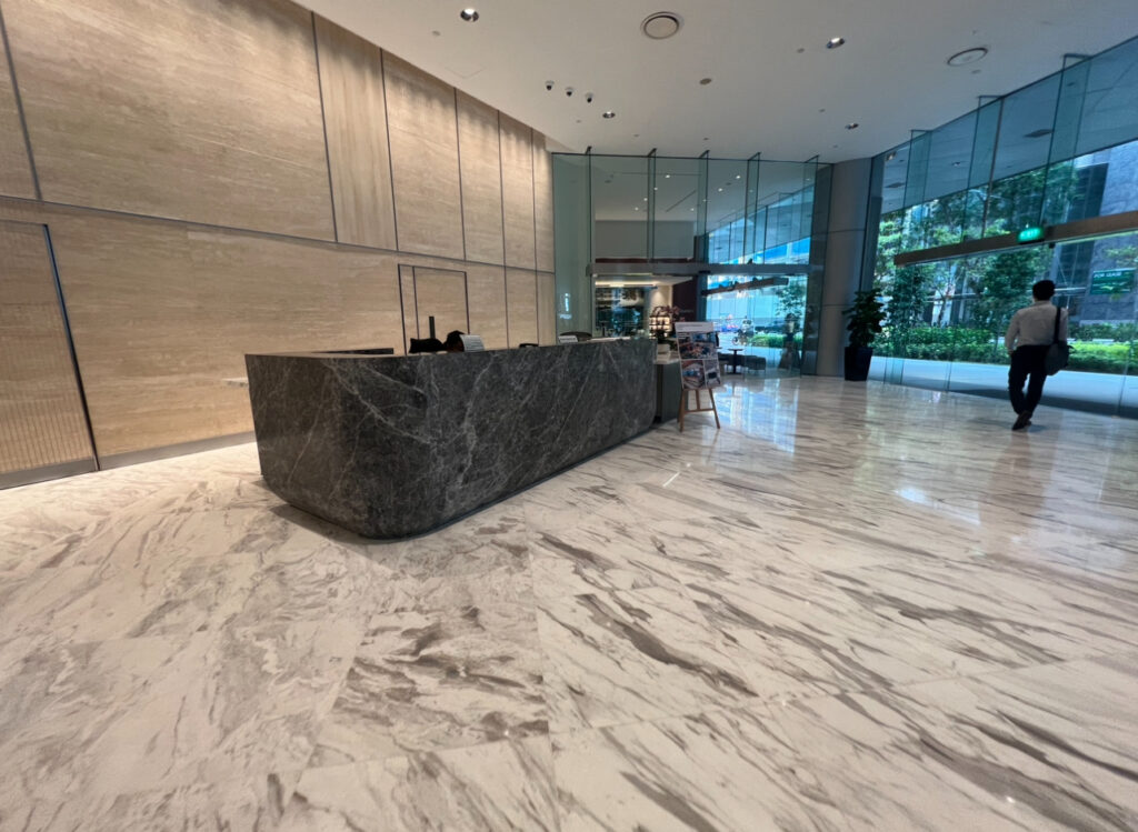 A modern office lobby with a marble floor, a large dark stone reception desk, glass walls, and a person walking towards the exit.