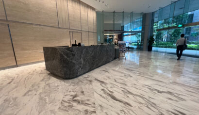 A modern office lobby with a marble floor, a large dark stone reception desk, glass walls, and a person walking towards the exit.