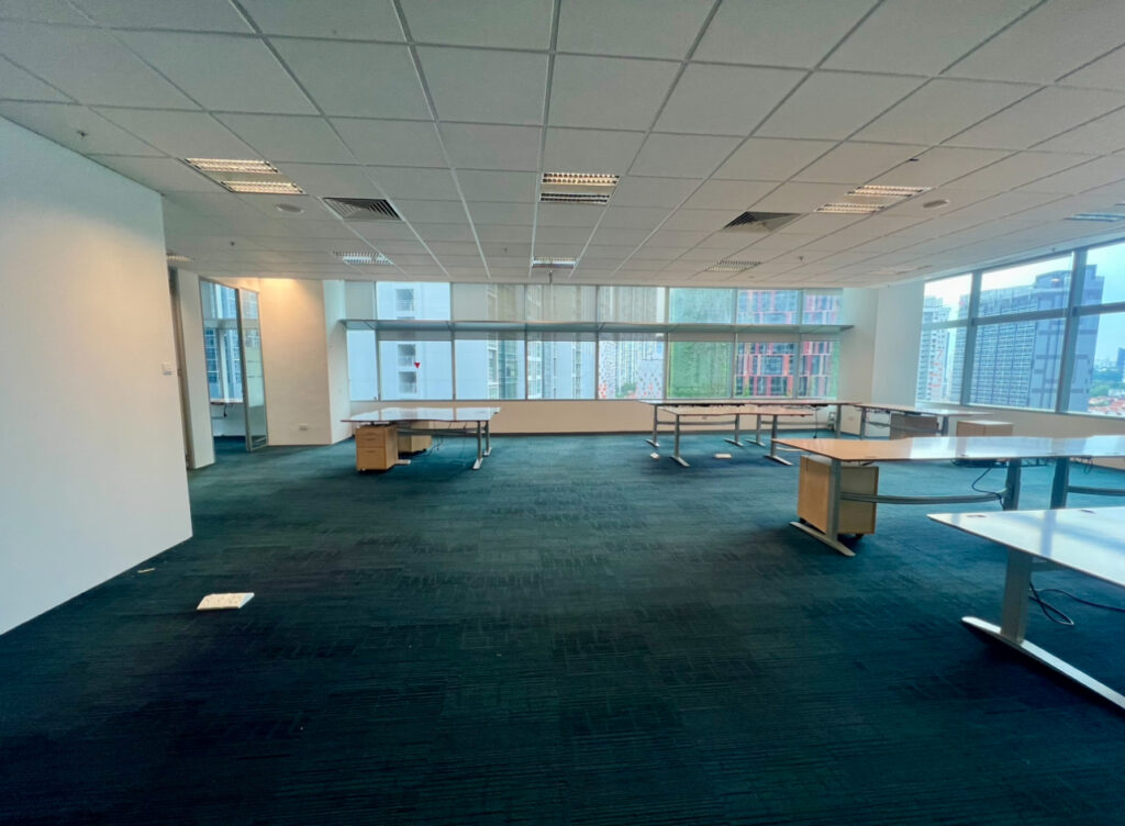 Spacious, empty office with several desks, carpeted floor, white walls, ceiling lights, and large windows overlooking city buildings outside.