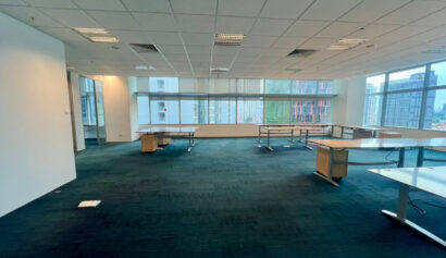 Spacious, empty office with several desks, carpeted floor, white walls, ceiling lights, and large windows overlooking city buildings outside.