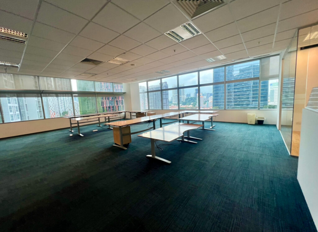 Spacious, empty office with several desks, large windows on one side, and a view of city buildings outside.