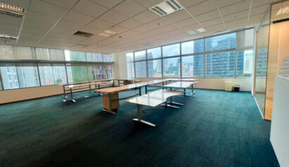 Spacious, empty office with several desks, large windows on one side, and a view of city buildings outside.