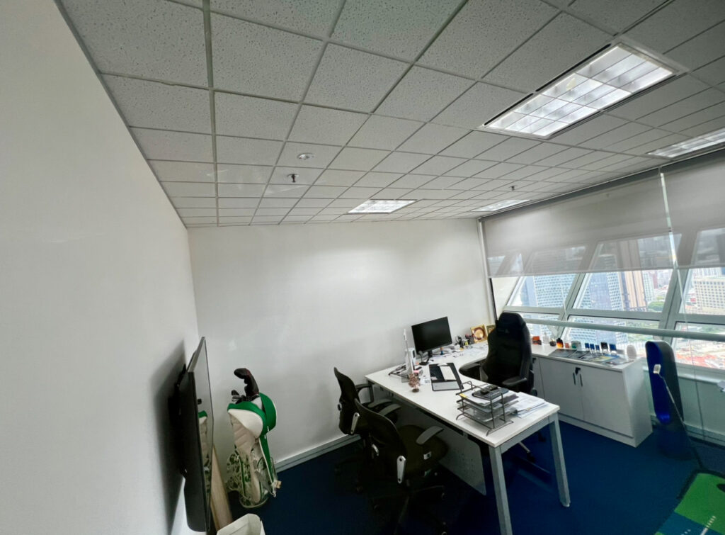 Modern office with two desks, computer monitors, office chairs, and a window offering a city view; a golf bag and TV are visible on the left side.