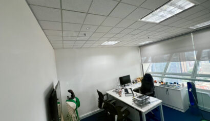 Modern office with two desks, computer monitors, office chairs, and a window offering a city view; a golf bag and TV are visible on the left side.