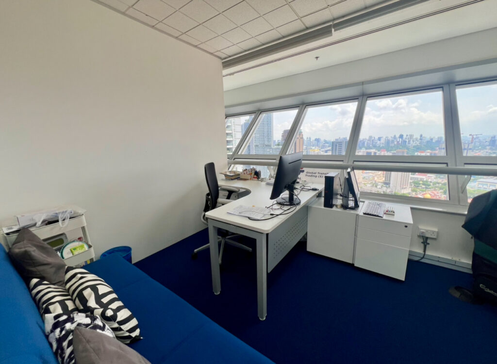 A modern office with a desk, computer, two chairs, a blue sofa with cushions, a small cupboard, and large windows overlooking a cityscape.