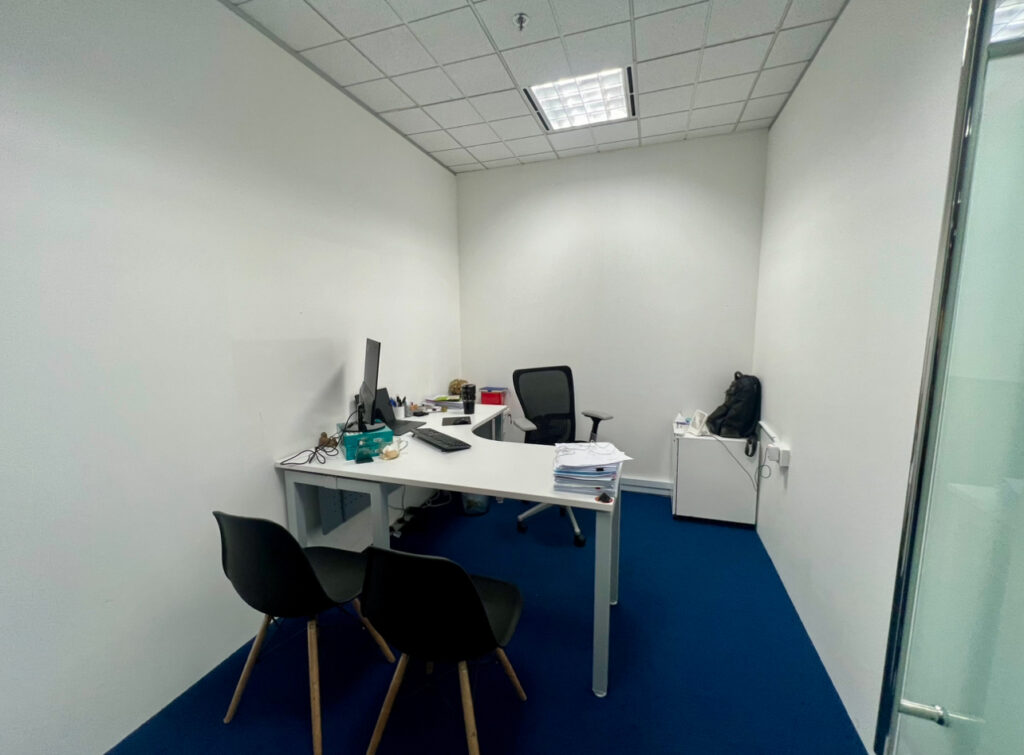 Small office with a white desk, computer monitor, office chair, two visitor chairs, and a side cupboard with a bag; blue carpet and white walls.
