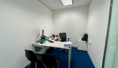 Small office with a white desk, computer monitor, office chair, two visitor chairs, and a side cupboard with a bag; blue carpet and white walls.
