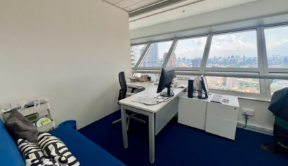 A modern office with a desk, computer, two chairs, a blue sofa with cushions, a small cupboard, and large windows overlooking a cityscape.
