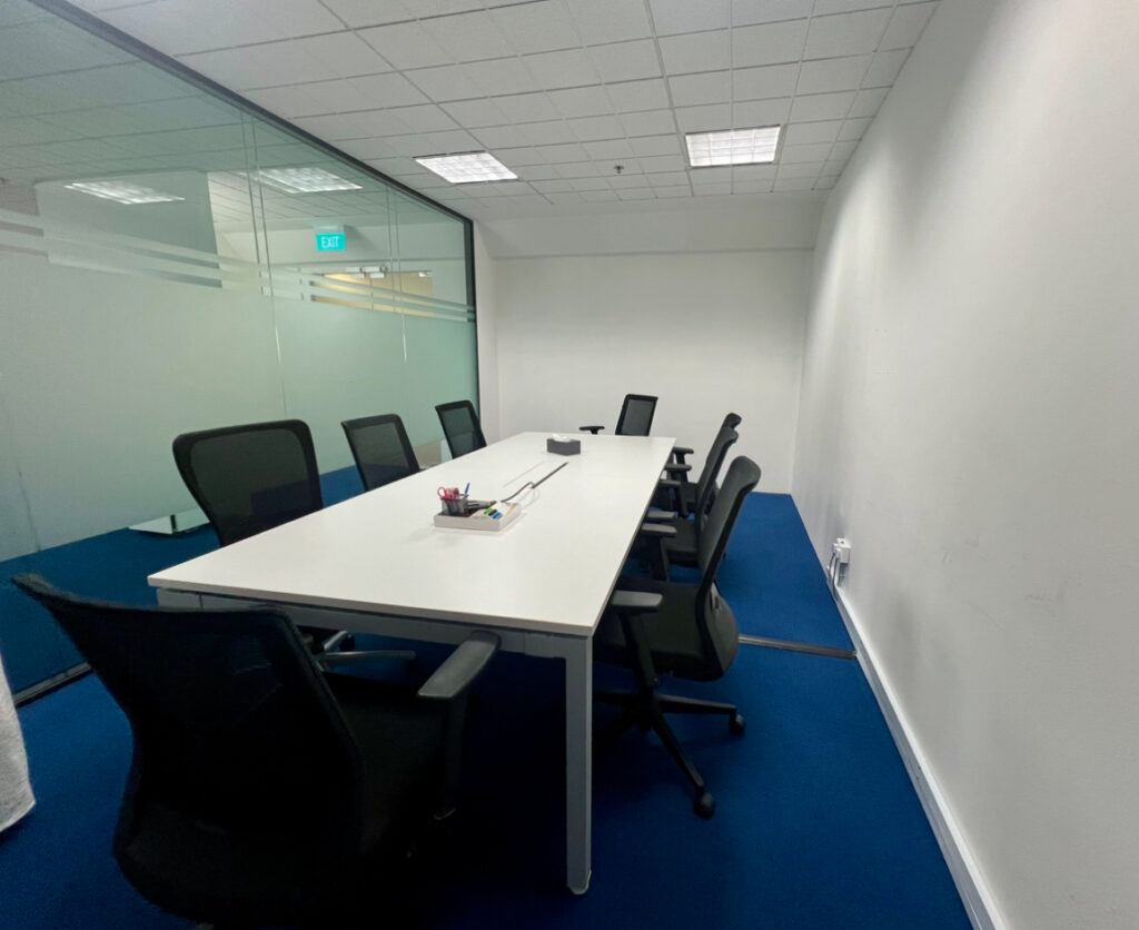 A modern conference room with a long white table, six black office chairs, blue carpet, and glass walls. Stationery items are on the table. Room is empty and well-lit.