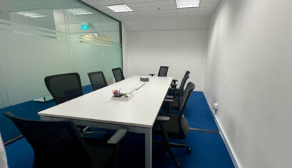 A modern conference room with a long white table, six black office chairs, blue carpet, and glass walls. Stationery items are on the table. Room is empty and well-lit.