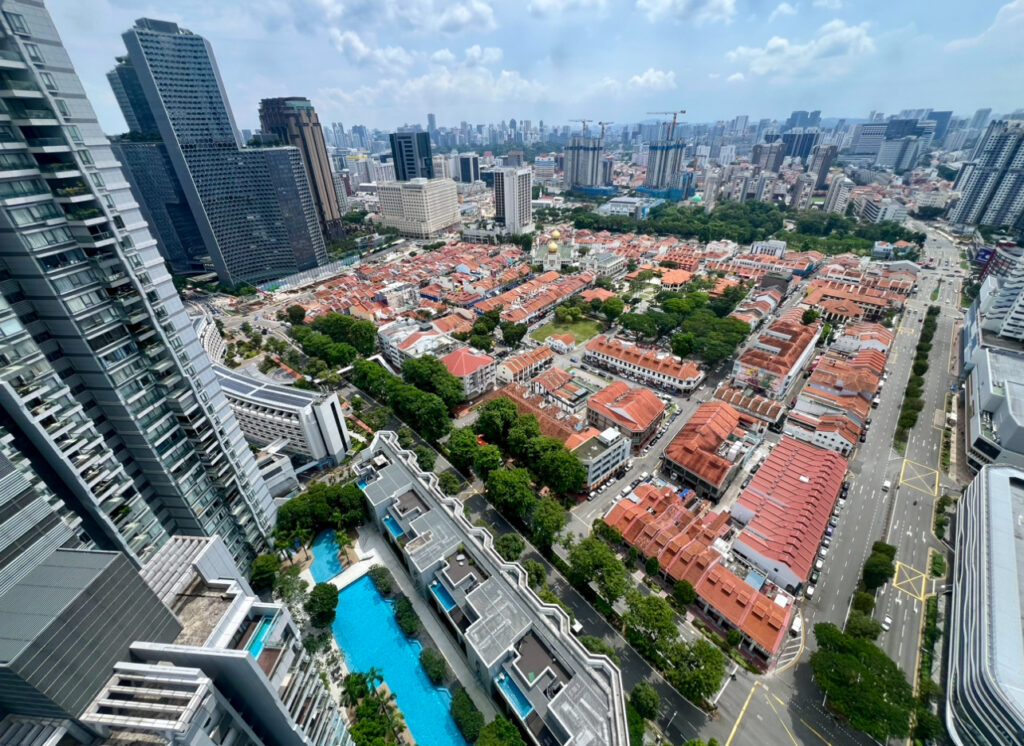 Aerial view of a city with high-rise buildings, red-roofed houses, tree-lined streets, and a blue swimming pool under a partly cloudy sky.