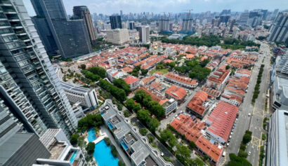 Aerial view of a city with high-rise buildings, red-roofed houses, tree-lined streets, and a blue swimming pool under a partly cloudy sky.
