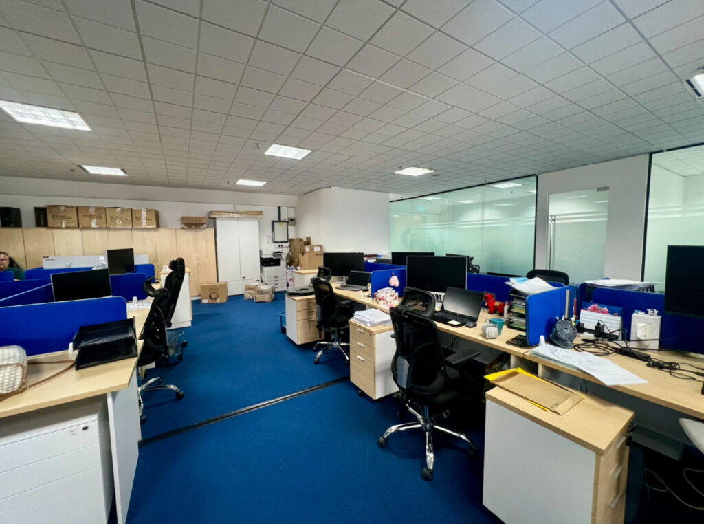Modern office with blue carpet, several desks and office chairs, desktop computers, and scattered office supplies. Cardboard boxes and shelves are visible in the background.