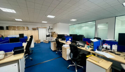 Modern office with blue carpet, several desks and office chairs, desktop computers, and scattered office supplies. Cardboard boxes and shelves are visible in the background.
