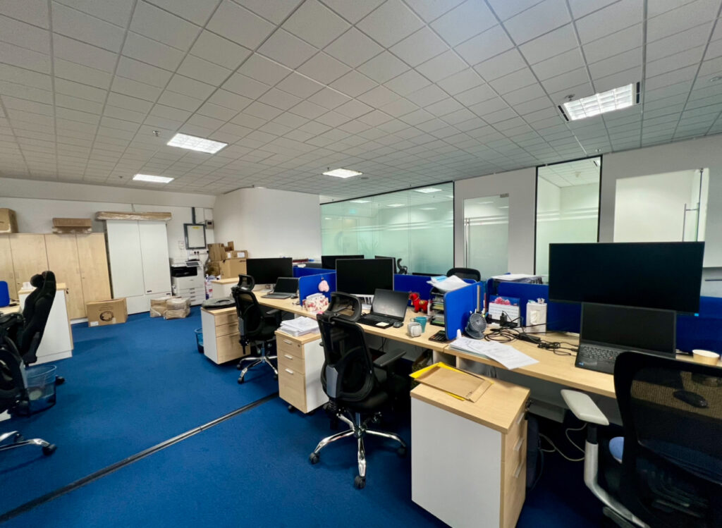 Modern office with blue carpet, several empty workstations, computer monitors, office chairs, and various office supplies scattered about. Cardboard boxes and cupboards are visible in the background.