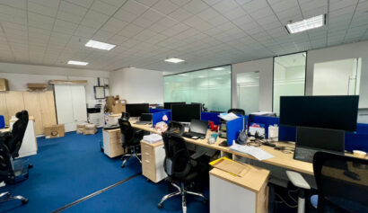 Modern office with blue carpet, several empty workstations, computer monitors, office chairs, and various office supplies scattered about. Cardboard boxes and cupboards are visible in the background.