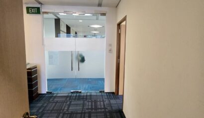 A corridor with patterned carpet leads to a glass door in an office setting. An exit sign is visible above the doorway on the left wall.