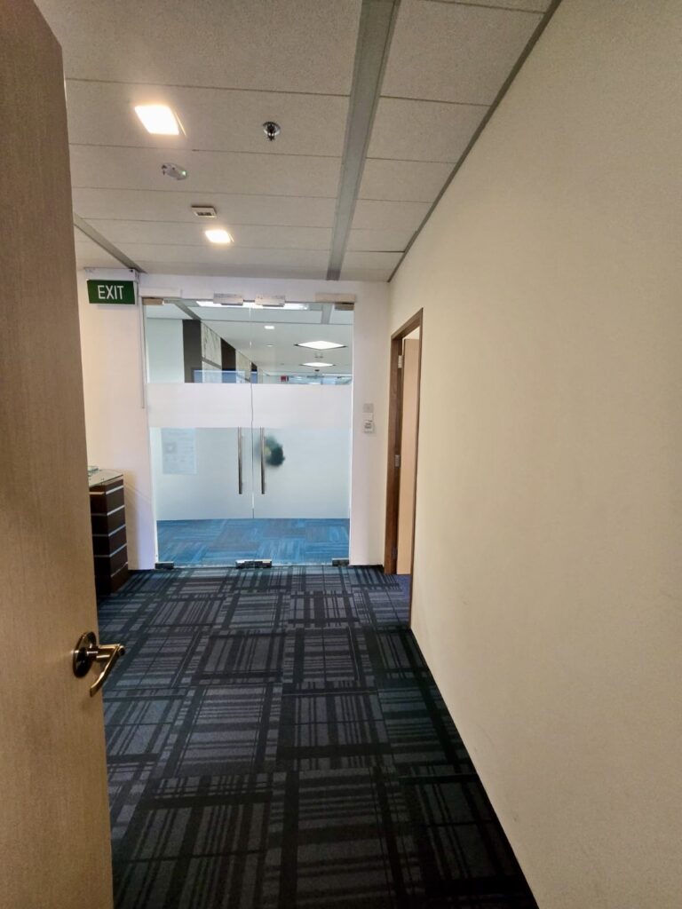 A corridor with patterned carpet leads to a glass door in an office setting. An exit sign is visible above the doorway on the left wall.