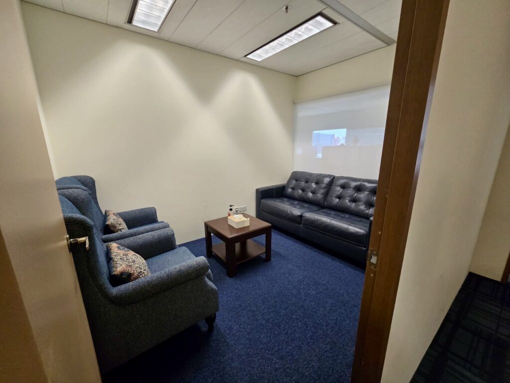 A small office room with two armchairs, a black sofa, a wooden coffee table with tissues, blue carpet, and bright overhead lights.