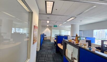 Office corridor with blue cubicles on the right, a glass window on the left, overhead lights, and large windows at the end of the room letting in daylight.
