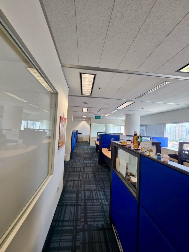 Office corridor with blue cubicles on the right, a glass window on the left, overhead lights, and large windows at the end of the room letting in daylight.