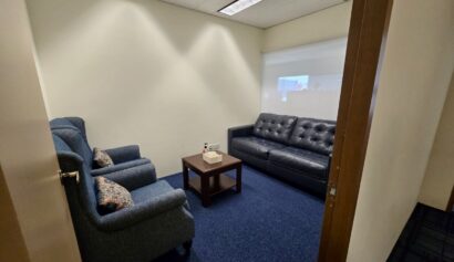 A small office room with two armchairs, a black sofa, a wooden coffee table with tissues, blue carpet, and bright overhead lights.