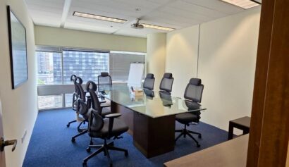 A modern conference room with a glass table, eight office chairs, a flip chart, and large windows overlooking city buildings.
