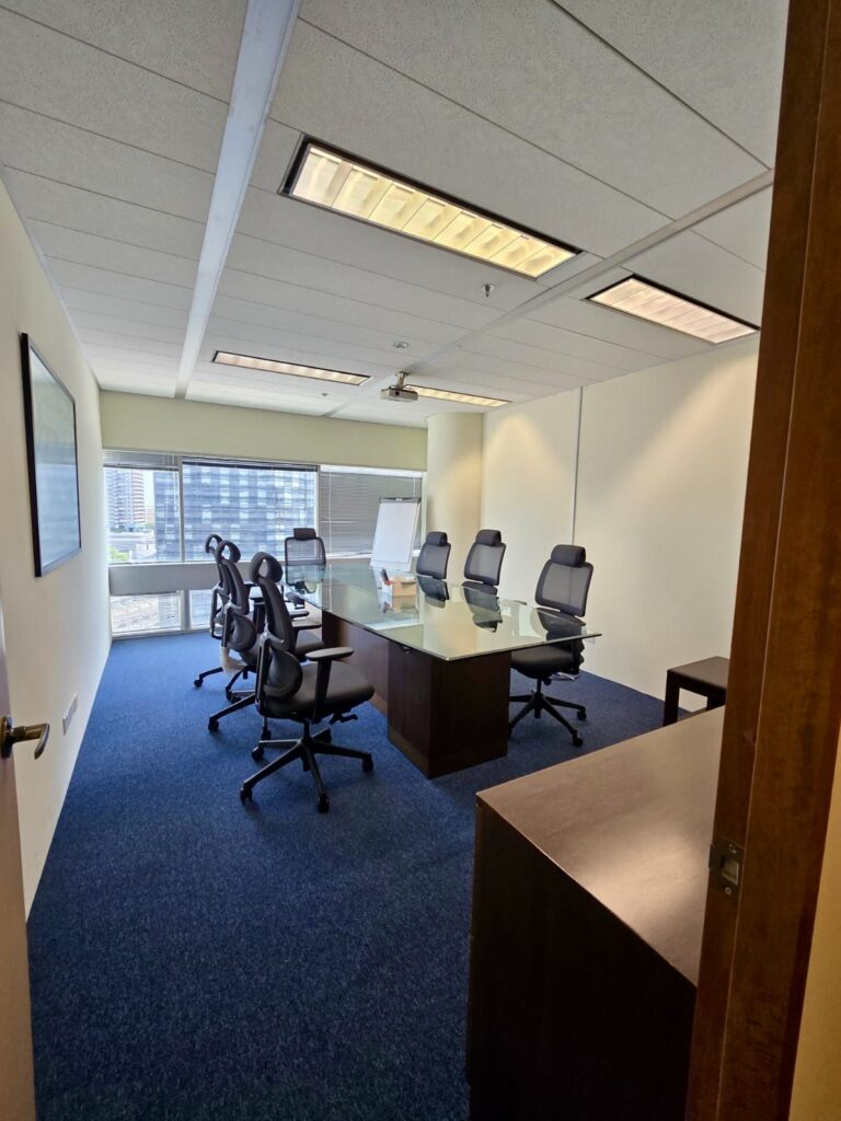 A modern conference room with a glass table, eight office chairs, a flip chart, and large windows overlooking city buildings.