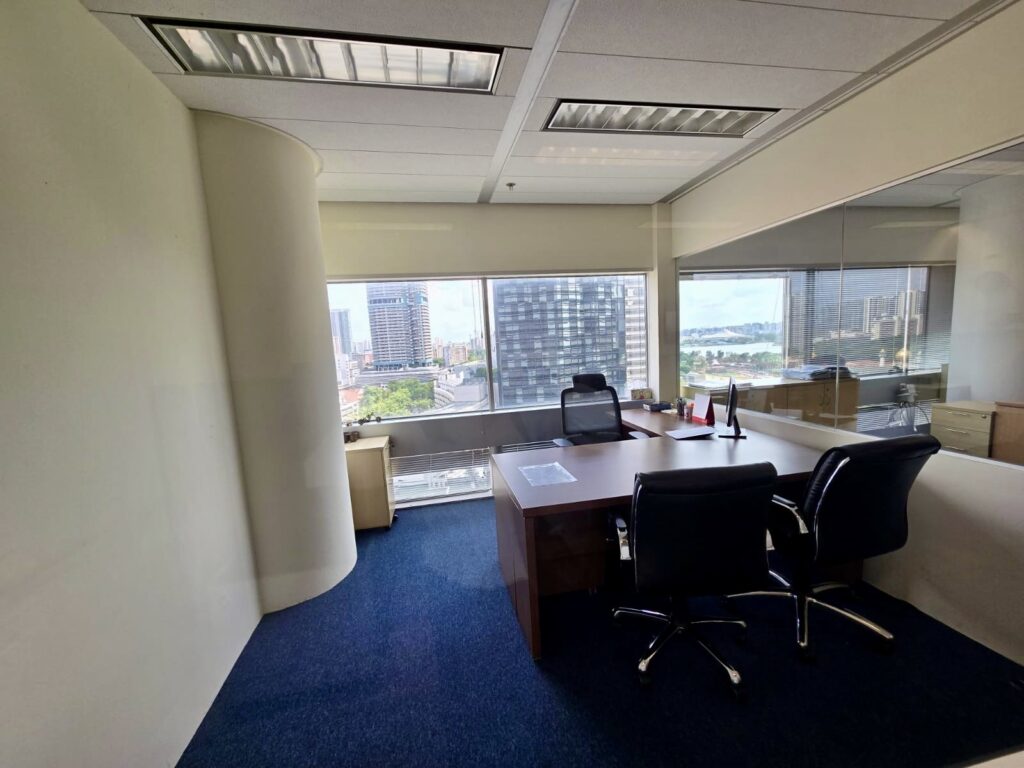 Modern office with two chairs, a large desk, and windows overlooking a cityscape and water. The room has white walls, blue carpet, and natural light.