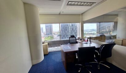 Modern office with two chairs, a large desk, and windows overlooking a cityscape and water. The room has white walls, blue carpet, and natural light.